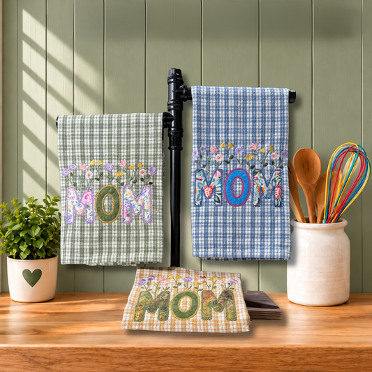 Three embroidered kitchen towels hanging on a black wall-mounted rack, with a folded matching towel in front on a wooden countertop. Sage green gingham, Golden yellow gingham, and Heritage blue gingham towels; each with a floral embroidery and the word 'MOM' appliqued underneath.