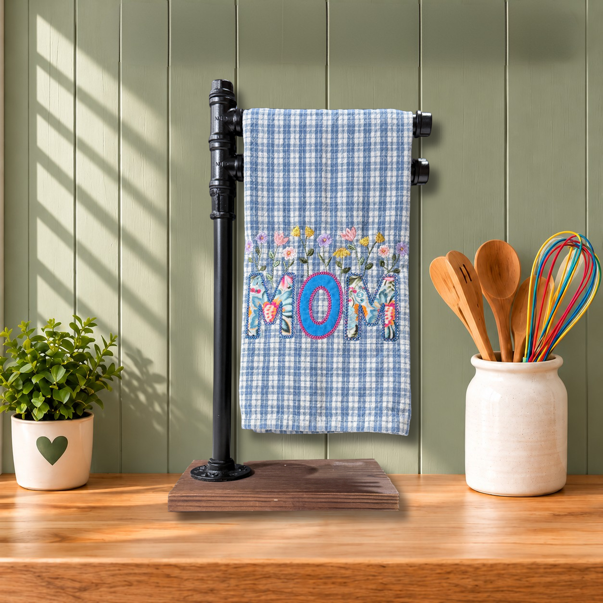 A kitchen towel with a Heritage blue gingham pattern and a colorful floral design spelling 'MOM'.