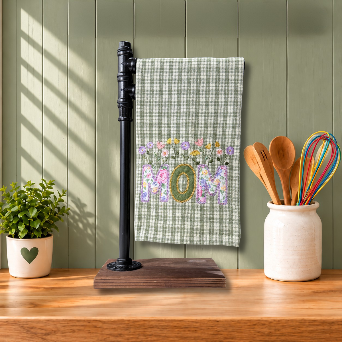 A kitchen towel with a Sage green gingham pattern and a colorful floral design spelling 'MOM'.
